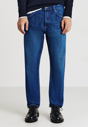 Jeans di denim blu con taglio dritto, cuciture visibili e design classico a cinque tasche, abbinati a scarpe nere con lacci.
