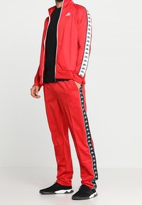 Roter Trainingsanzug aus glattem Stoff, mit weißem Reißverschluss und seitlichen Streifen sowie schwarzen Logo-Akzenten. Kombination mit schwarzen Sportschuhen.