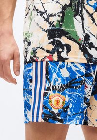 Χέρι που φοράει σορτς με μπλε, μαύρο, λευκό και μπεζ αφηρημένο σχέδιο και έμβλημα της Μάντσεστερ Γιουνάιτεντ στον μηρό με κάθετες ρίγες.