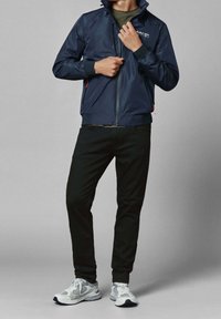 Mies, joka pukeutuu tummansiniseen Red Bull Racing -takkiin, oliivinvihreään paitaan, mustiin housuihin ja valko-harmaisiin lenkkareihin, säätää takin vetoketjua.