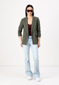 Sacou verde măsliniu cu o croială ajustată, top burgundy cu decolteu adânc în V, blugi deschiși la culoare, evazați, și adidași albi. Ochelari de soare mari, maro, incluși.