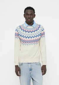 Μπεζ πουλόβερ με πολύχρωμο γεωμετρικό μοτίβο σε παστέλ ροζ, μπλε και ανοιχτό μπλε. Με στρογγυλή λαιμόκοψη και ριμπ μανσέτες, φορεμένο πάνω από μπλε πουκάμισο με γιακά.