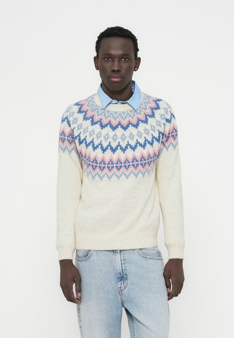Μπεζ πουλόβερ με πολύχρωμο γεωμετρικό μοτίβο σε παστέλ ροζ, μπλε και ανοιχτό μπλε. Με στρογγυλή λαιμόκοψη και ριμπ μανσέτες, φορεμένο πάνω από μπλε πουκάμισο με γιακά.