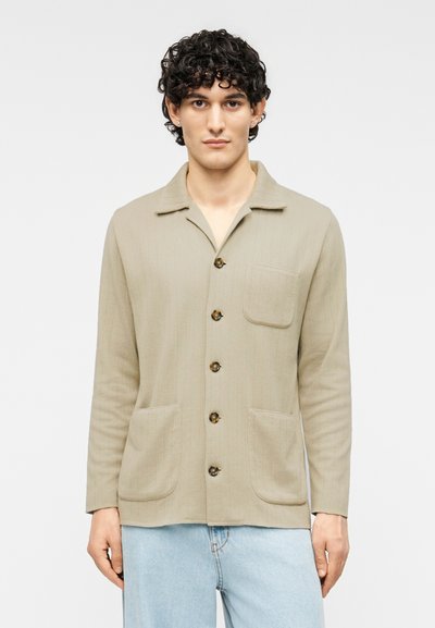 Light beige button-up jacket with vertical ribbing, a collar, a chest pocket, two front pockets, and brown buttons. Worn over blue jeans.