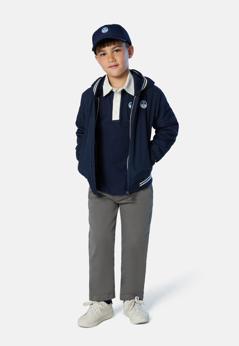 Giubbotto bomber blu navy con rifiniture bianche, polo blu navy, pantaloni grigi, scarpe da ginnastica bianche e cappellino blu navy con logo. Ragazzo giovane in piedi con le mani nelle tasche.