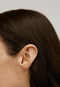 Pendiente de oro con diseño delicado de pájaro, adornado con pequeñas gemas transparentes. El cuff se lleva en la oreja izquierda, contra el cabello oscuro.