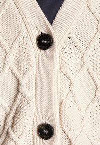 Cárdigan de punto beige con un patrón de trenzas, dos botones negros y un diseño de escote en V. La textura parece suave y gruesa.