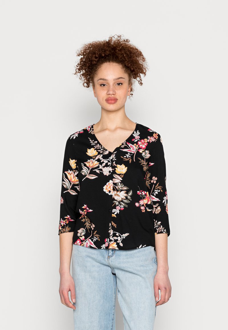 Blusa preta com estampado floral, com mangas 3/4, apresentando padrões coloridos de flores, um decote em V e tecido leve e suave. Combinada com jeans azuis.