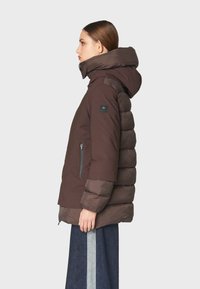 Chaqueta acolchada marrón con un alto cuello y capucha, que cuenta con un bolsillo lateral con cremallera y un parche de logo en la manga. Textura suave y diseño en capas.