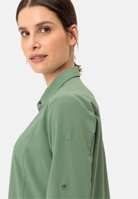 Camicia verde con colletto, caratterizzata da una texture liscia, maniche lunghe e un dettaglio decorativo ricamato in verde sulla manica sinistra.