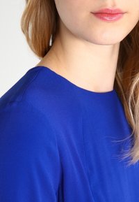 Królestwo niebieski top o gładkiej, matowej fakturze i okrągłym dekolcie. Zbliżenie uwydatnia detale ramion i włosów.