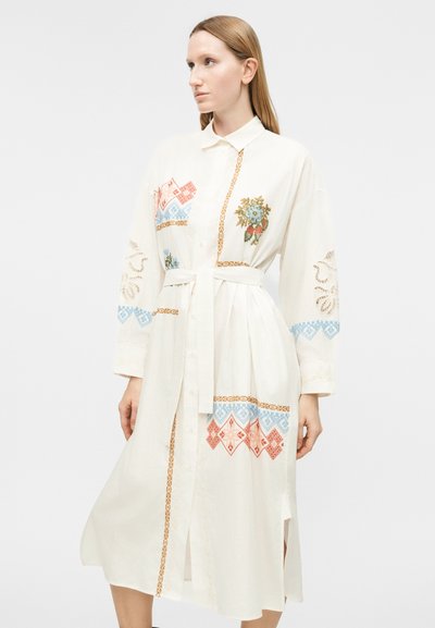 Γυναίκα που φοράει λευκό φόρεμα μέσης μήκους με γιακά, ζώνη με φιόγκο και πολύχρωμα κεντήματα σε μπλε, κόκκινο και καφέ, στέκεται μπροστά σε απλό φόντο.