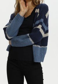 Kvinna iförd en blå och marinrandig stickad cardigan över en svart topp och svarta byxor, poserande med en hand nära sitt ansikte.