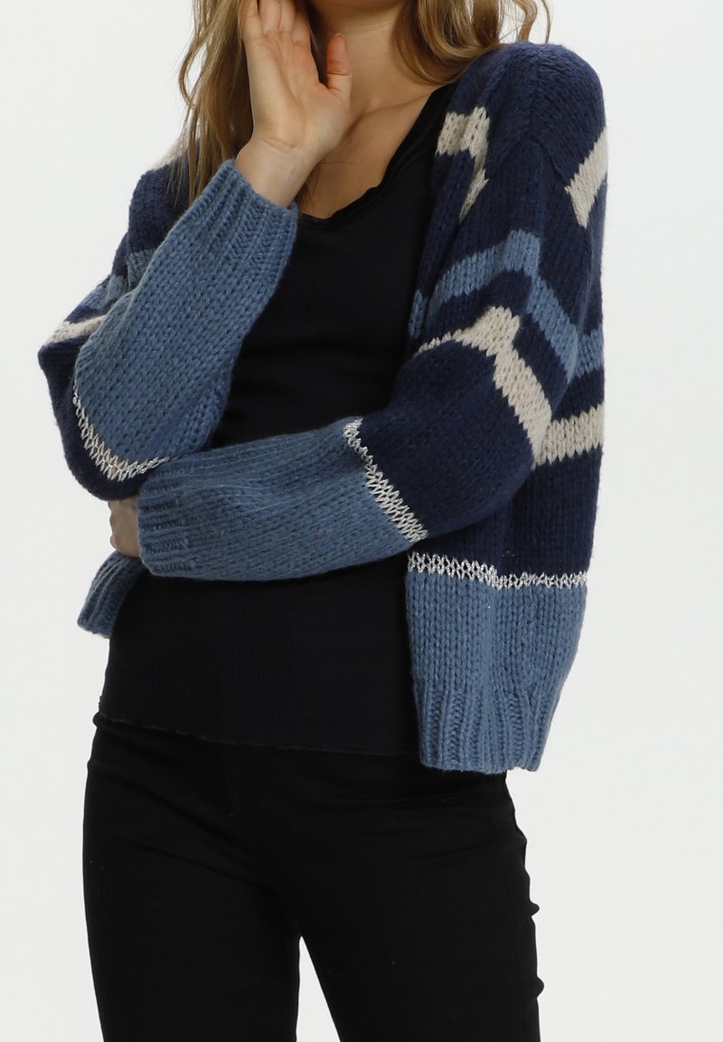 Kvinna iförd en blå och marinrandig stickad cardigan över en svart topp och svarta byxor, poserande med en hand nära sitt ansikte.