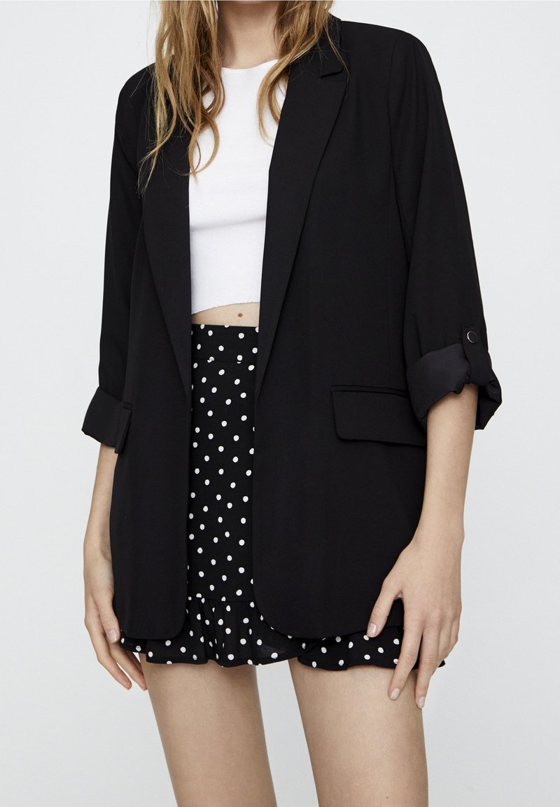 Femme portant un blazer noir aux manches retroussées sur un crop top blanc et une mini-jupe noire à pois avec un ourlet froufroutant sur fond uni.