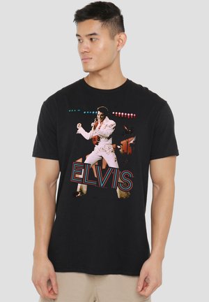 Young man wearing black t-shirt featuring Elvis Presley in white jumpsuit singing on stage with bright lights and large "ELVIS" text.