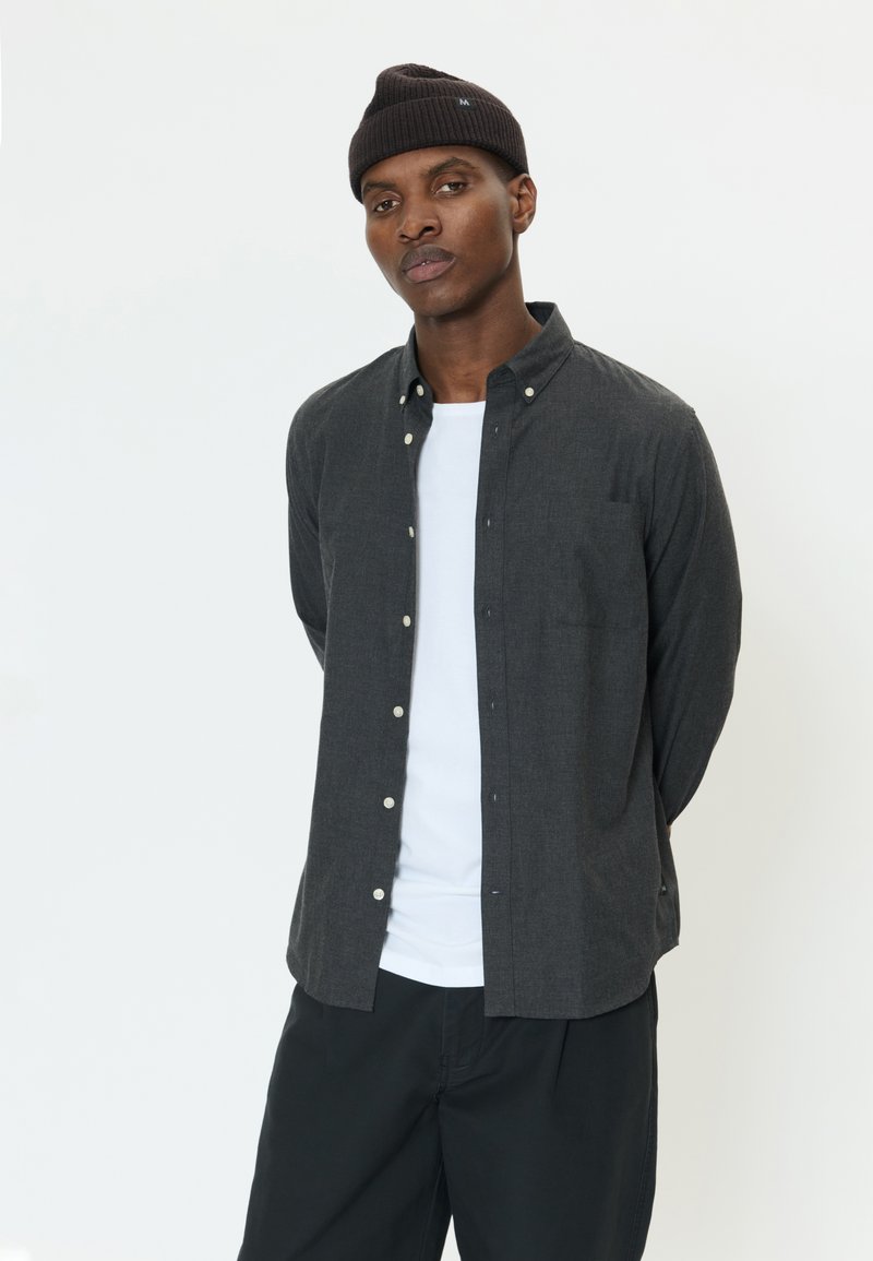 Hombre que lleva una camisa oscura abotonada sobre una camiseta blanca, pantalones negros y un gorro oscuro, de pie con las manos detrás de la espalda.