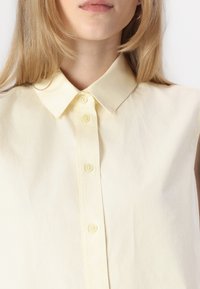 Camisa sin mangas amarilla con cuello clásico y cierre de botones en el frente, confeccionada con una tela ligera y arrugada.