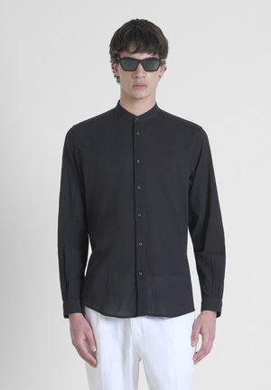 Camicetta nera a maniche lunghe con texture verticale, colletto alla coreana e chiusura frontale con bottoni. Indossata con pantaloni bianchi e occhiali da sole scuri.