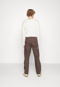 Camisa de manga larga color crema con el texto "NIGHT ADDICT", combinada con pantalones cargo marrones que tienen bolsillos laterales y un pequeño parche de logo.
