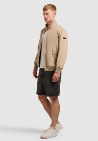 Homme portant une veste zippée beige, un t-shirt blanc, un short en jean noir et des baskets blanches, debout devant un fond uni.