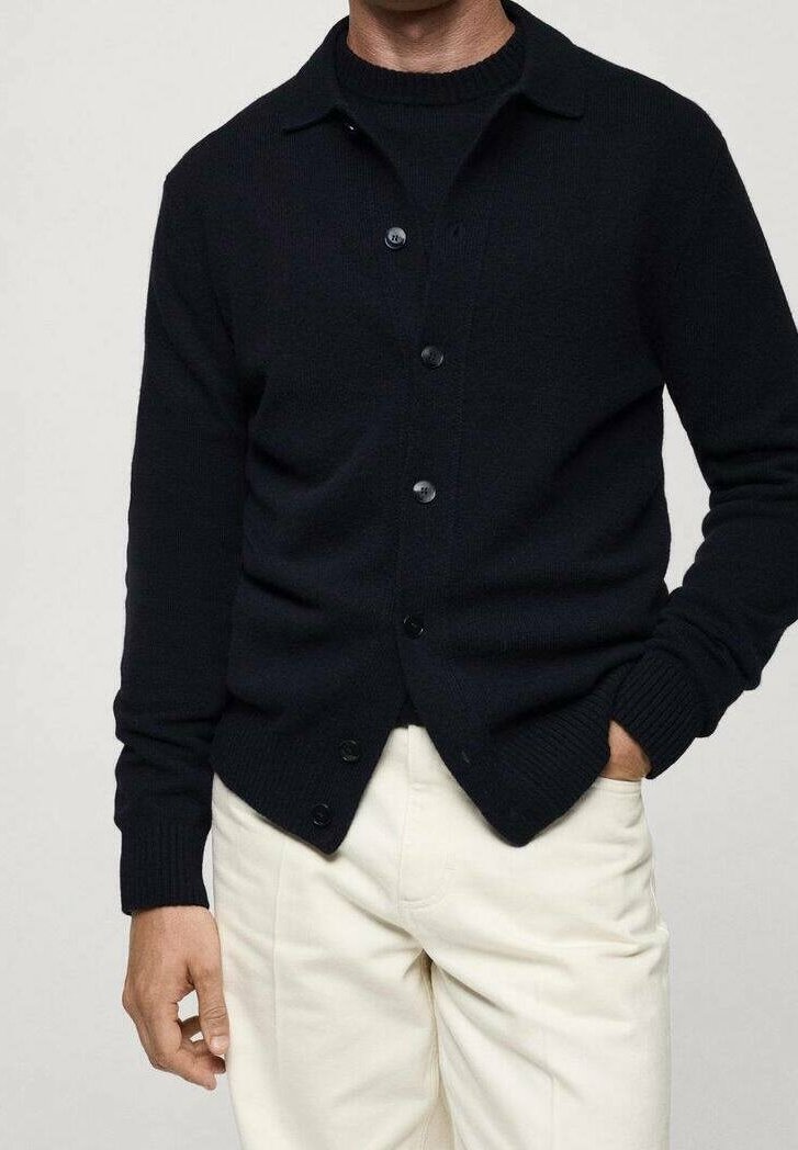 Schwarzer, gestrickter Strickjacke mit Knopfleiste und Kragen, ausgestattet mit gerippten Bündchen und Saum, getragen über einem schwarzen Pullover und cremefarbenen Hosen.