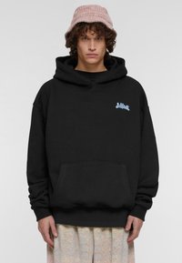 Sudadera negra con capucha y bolsillo delantero, con un logo azul en el pecho, combinada con un sombrero tipo cubo de color rosa claro.