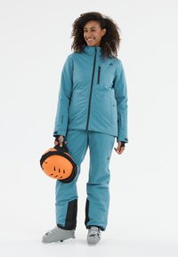 Blaue Skijacke und Hose, beide aus strukturiertem Material gefertigt. Merkmale sind eine Reißverschlusstasche, ein oranger Helm und graue Skischuhe.