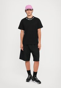 Svart T-skjorte og shorts, rosa caps, svarte sokker og svarte joggesko. T-skjorten har hvit tekst på kragen.
