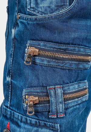 Denim broek met meerdere ritsen in bronzen hardware, in een vervaagde blauwe kleur, contrasterende rode stiksels en een robuuste textuur.