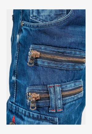 Pantaloni in denim con numerosi zip in metallo bronzo, di colore blu sbiadito, con cuciture rosse a contrasto e una texture robusta.
