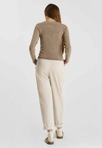Femme debout de dos, portant un haut marron moulant à manches longues, un pantalon ample couleur crème, des chaussettes à motifs et des sandales beiges.