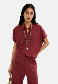 Koszula na krótki rękaw w kolorze burgundowym, zapinana na guziki, wykonana z miękkiego materiału, w zestawieniu z pasującymi spodniami. Uzupełniona warstwowymi naszyjnikami i torebką.