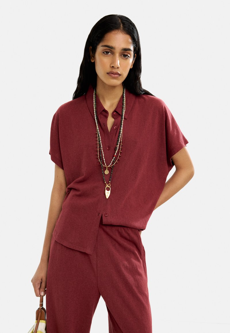 Koszula na krótki rękaw w kolorze burgundowym, zapinana na guziki, wykonana z miękkiego materiału, w zestawieniu z pasującymi spodniami. Uzupełniona warstwowymi naszyjnikami i torebką.