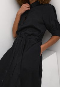 Robe chemise noire avec un devant à boutons, des manches courtes bouffantes et une taille nouée. Présente une texture lisse et des poches latérales.