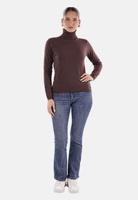 Maglione a collo alto marrone realizzato in tessuto morbido, abbinato a jeans blu. L'outfit si completa con sneakers bianche, creando un look casual.