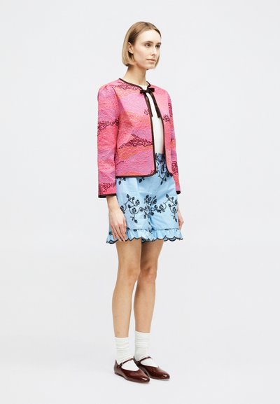 Γυναίκα στέκεται φορώντας ένα ροζ σακάκι με σχέδια, γαλάζια κεντημένα σορτς με βολάν στο κάτω μέρος, λευκές κάλτσες και μπορντό παπούτσια Mary Jane.