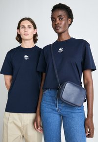 Dwie osoby noszą granatowe bawełniane koszulki z białym logo Lacoste. Kobieta trzyma małą granatową skórzaną torebkę, w zestawieniu z niebieskimi dżinsami.
