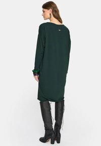 Kobieta ubrana w ciemnozieloną, długą sukienkę midi z długim rękawem, czarne, wiązane kozaki za kolano oraz masywne bransoletki, stoi plecami do białego tła.