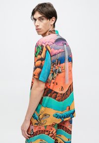 Πουκάμισο με κοντά μανίκια που παρουσιάζει πολύχρωμο σχέδιο ερήμου, με έντονες αποχρώσεις πορτοκαλί, μπλε και πράσινου· κατασκευασμένο από ελαφρύ ύφασμα.