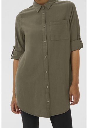 Femme portant une chemise boutonnée vert olive avec une poche poitrine et des manches retroussées, associée à un pantalon noir.