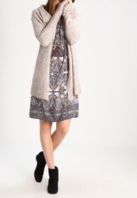 Kobieta stojąca z skrzyżowanymi nogami, ubrana w wzorzystą sukienkę do kolan, beżowy kardigan i czarne botki za kostkę na białym tle.