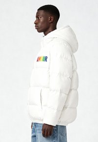 Doudoune blanche avec capuche, arborant une broderie colorée "AMOUR". Tissu doux, design matelassé et poches avant.