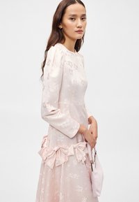 Ανοιχτό ροζ φόρεμα με floral κέντημα, μακρύ μανίκι και μώβ λεπτομέρεια στη μέση. Το μοντέλο κρατά μια μικρή ανοιχτό ροζ τσάντα.