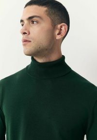 Pulover de culoare verde închis, cu guler tip turta, confecționat dintr-un material moale și ușor. Are un gât texturat și o textură netedă, cu un design ajustat.