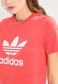 Korálové bavlněné tričko s výrazným bílým logem Adidas a designem trojlístku a třemi vodorovnými pruhy. Krátké rukávy, kulatý výstřih.