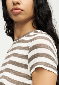 Νεαρή γυναίκα με μακριά σκούρα μαλλιά, που φοράει μια κοντομάνικη λευκή και ανοιχτοκάστανη πλεκτή μπλούζα με οριζόντιες ρίγες, εμφανιζόμενη από τους ώμους και πάνω.