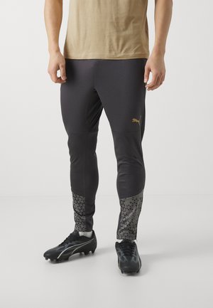 Zwarte sportbroek met gestructureerde grijze onderstukken met een driehoekig patroon. Gekleed met zwarte sportschoenen en een beige shirt.