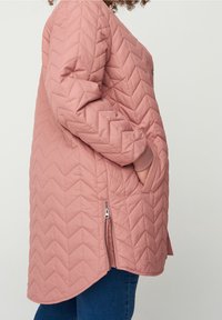 Růžový prošívaný kabát s vzorem v šagénovém stylu, bočními kapsami a zipem na spodním okraji. Má zaoblený lem a žebrované manžety.