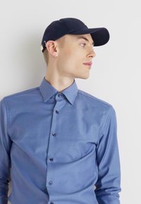 Button-up shirt in light blue with subtle vertical stripes, long sleeves, and a pointed collar, paired with a black cap.